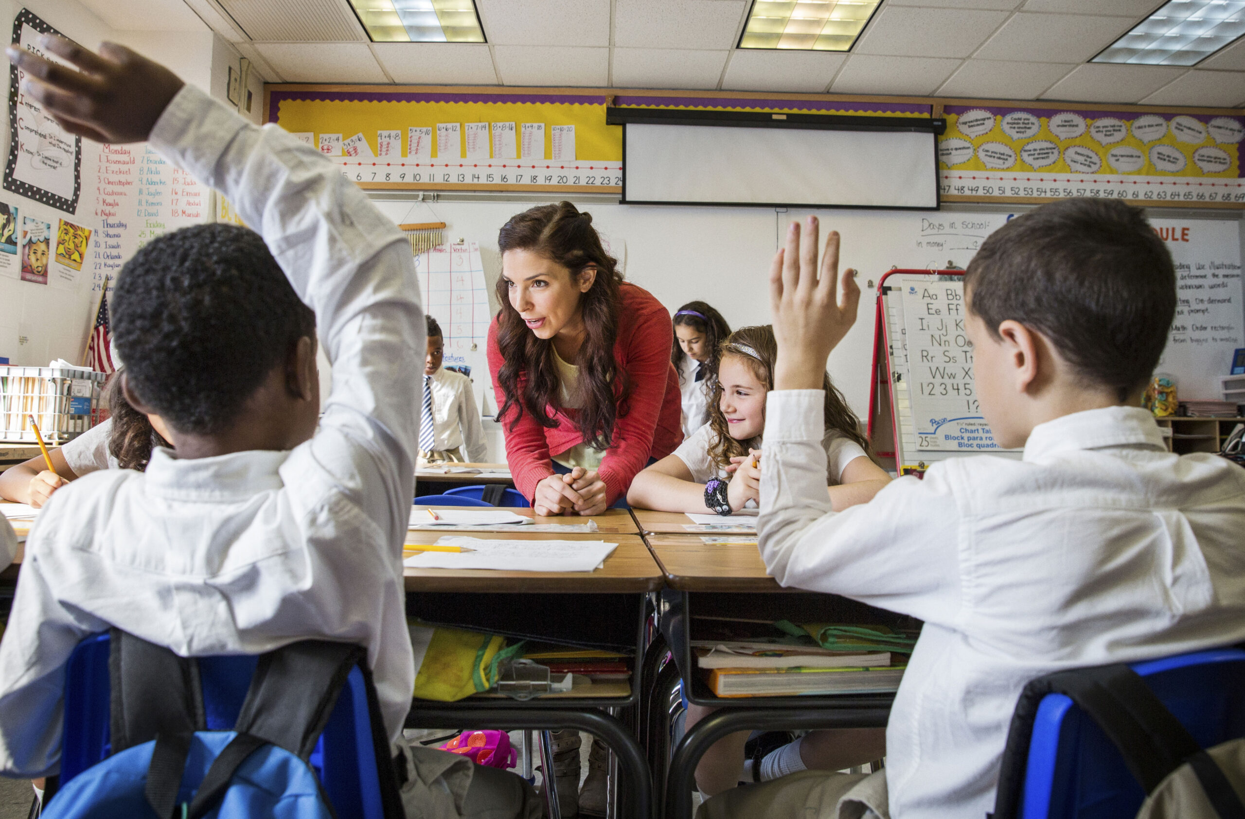 female teacher in classroom with two schoolboys (8-9 and 10-11) raising hands in foreground