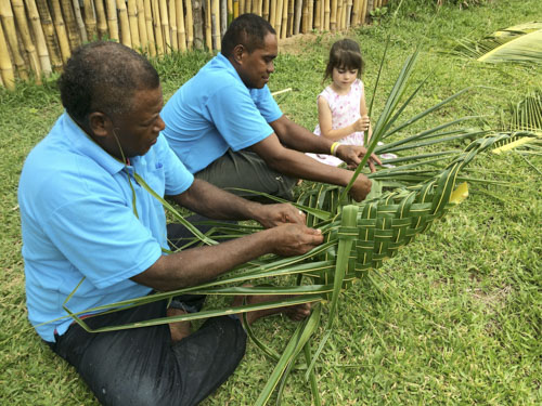 Fijian men teaching a young girl how to weave a basket.
